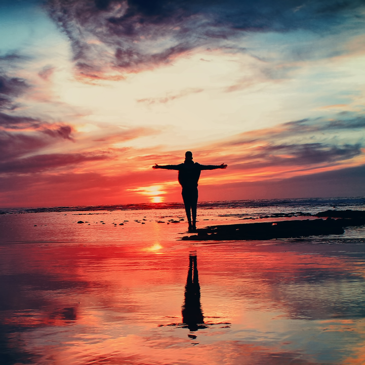 Picture of a figure with arms outstretched on a beach against a vivid red and orange sunset.